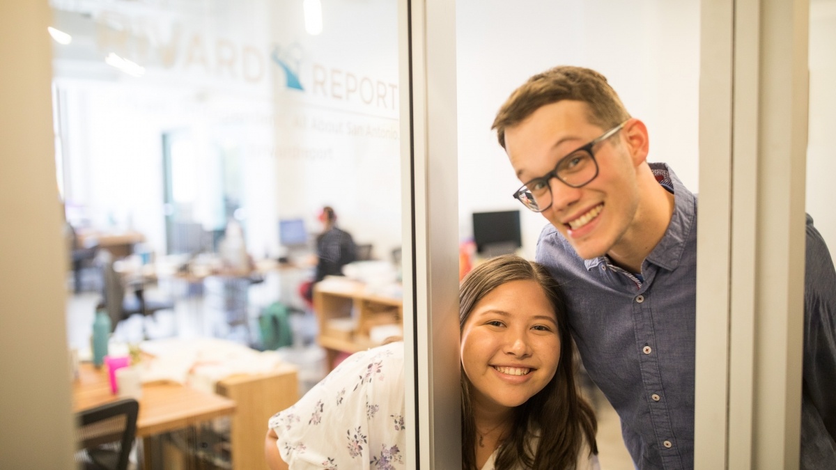 Two students peaking through glass door at Rivard Report