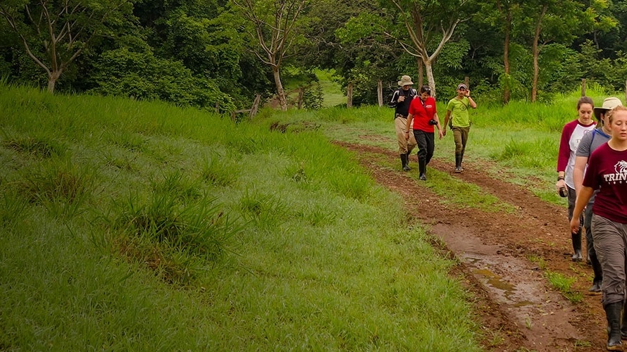 students hike along a muddy path in a lush field
